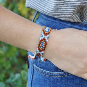 May include: A woven friendship bracelet featuring a geometric diamond pattern in shades of orange, blue, and white. The bracelet is constructed from thin threads arranged in a repeating symmetrical design that wraps around the wrist.