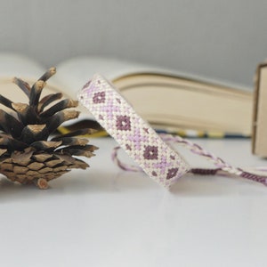 以下が含まれることがあります： ラベンダーとクリーム色の幾何学模様が特徴の手作りのフレンドシップブレスレット。 ブレスレットは織り糸で作られており、繰り返しのダイヤモンドデザインが特徴です。 ブレスレットは白い表面に表示されています。