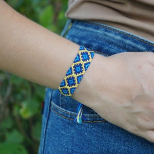 May include: A woven friendship bracelet featuring a repeating diamond pattern in shades of bright yellow, royal blue, and light blue. The band is constructed from knotted thread and includes small dangling fringe ends at the closure point.