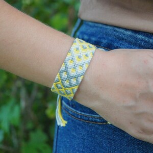 May include: A woven friendship bracelet featuring a geometric pattern in shades of yellow, grey, and white. The bracelet has a wide, flat band design with a repeating diamond-like motif and includes small, dangling thread tassels at the end.