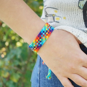 May include: A woven friendship bracelet featuring a vibrant, multi-coloured geometric pattern in shades of blue, red, yellow, orange, and purple. The bracelet is constructed from thin thread and includes dangling braided ties at the end.