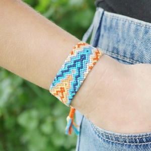 May include: A woven friendship bracelet in shades of blue, orange, and white, featuring a zig-zag pattern. The bracelet is worn on a wrist, with denim jeans and a black top in the background.