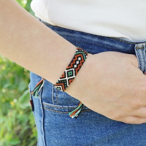 May include: A woven friendship bracelet featuring a geometric pattern in black, white, orange, and green thread. The bracelet has a flat, band-like construction with visible knotted ends and a repeating diamond and chevron design throughout the piece.