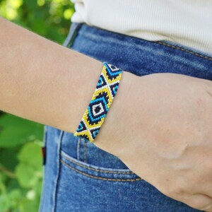 May include: A woven friendship bracelet featuring a repeating geometric diamond pattern in vibrant shades of navy blue, bright yellow, white, and light blue, constructed from multiple strands of thread in a flat, knotted design.
