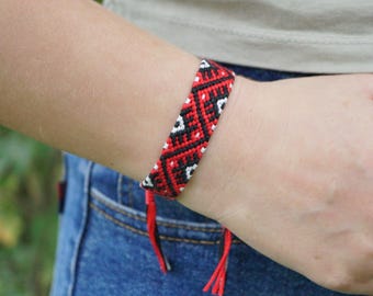 May include: A woven friendship bracelet featuring a geometric pattern in black, red, and white thread. The bracelet has a flat, wide band design with two loose, knotted thread ends hanging down from the bottom edge of the woven section.