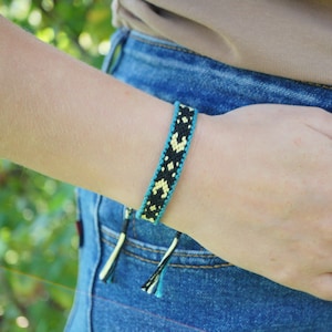 May include: A woven friendship bracelet featuring a repeating geometric pattern in black and pale yellow, bordered by thin teal blue lines. The bracelet is constructed from thread and includes two dangling tie ends at the bottom of the wristband.