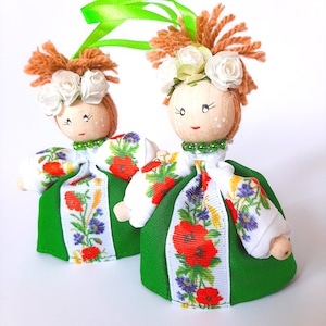 May include: Two handmade dolls dressed in traditional Ukrainian clothing. The dolls are wearing white shirts with red poppy embroidery, green skirts, and white floral wreaths.