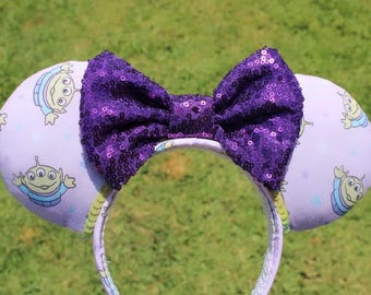 Puede incluir: Una diadema con dos orejas redondas cubiertas de tela color lavanda claro con un patrón repetitivo de alienígenas verdes de tres ojos con uniformes azules. Un gran lazo de lentejuelas de color morado oscuro está centrado entre las orejas.