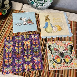 May include: Four different paper napkins with various designs. One napkin features a cartoon dog with a wheelbarrow, another has a rabbit with the text "Happy Easter", a third has a purple background with colorful butterflies, and the last one has a single butterfly with floral details on a beige background.