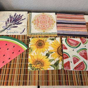 May include: A collection of decorative paper napkins with various designs. The napkins feature images of lavender, sunflowers, watermelon slices, and a striped pattern in shades of orange, red, and blue. The lavender napkin has the word "Lavendula".