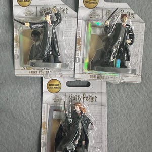 May include: Three Harry Potter character figurines in their original packaging. The figurines are of Harry Potter, Ron Weasley, and Hermione Granger. Each figure is holding a wand and wearing a black robe. The packaging includes the text "Harry Potter" and "Bonus Card".