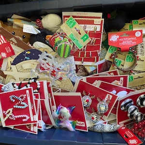 May include: A blue plastic storage bin filled with a variety of Christmas craft supplies, including red and white candy cane ornaments, a wooden reindeer, a silver glitter garland, and a mini bell garland. The bin also contains a variety of other Christmas decorations, such as wooden words, a wooden star, and a wooden Christmas tree.