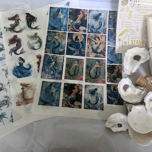 May include: A collection of mermaid-themed craft supplies. Includes sheets of mermaid images, oyster shells, small wooden boxes, a paintbrush, and decorative paper with nautical designs. The color palette features blues, whites, and natural tones.