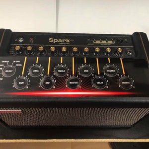 May include: Black guitar amplifier with gold knobs and the text "Spark" on the front. The amplifier has a black speaker grill and a red illuminated panel with control labels like "Gain", "Bass", and "Treble".
