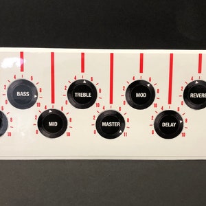 Könnte beinhalten: Weißes Bedienfeld mit schwarzen Knöpfen und roten Markierungen für einen Audioverstärker. Das Panel verfügt über Bedienelemente mit der Bezeichnung "Bass", "Treble", "Mod", "Reverb", "Delay" und "Output". Zusätzliche Bedienelemente sind "Select", "Gain" und "Mid".