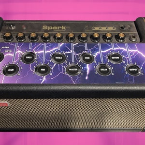 Peut inclure: Un amplificateur de guitare électrique noir avec une garniture dorée et un motif d'éclairs violet et noir. L'amplificateur possède une variété de boutons et de molettes, dont une étiquette "Spark 40".