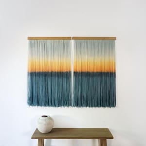 May include: Two ombre yarn wall hangings with a gradient of white, yellow, orange, and blue. The hangings are mounted on a wooden dowel rod.
