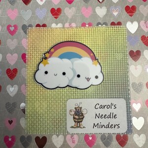 May include: A needle minder with a cartoon rainbow and cloud design. The needle minder is on a square of light green fabric, with a label that reads "Carol's Needle Minders" and a cartoon bee illustration. The background is covered in heart stickers.