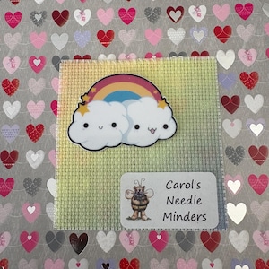 May include: A needle minder featuring a rainbow and two smiling cloud characters. The minder is set on a light green cross-stitch fabric, with a backdrop of hearts in shades of red, pink, and grey. The text "Carol's Needle Minders" is present.