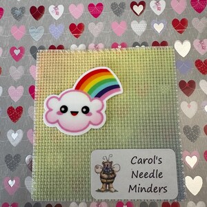 May include: A colourful rainbow cloud needle minder with a smiling face, placed on a green cross-stitch canvas. The image includes a label with a cartoon bee and the text "Carol's Needle Minders". The background is a silver surface with heart shapes.