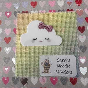 May include: A white cloud-shaped needle minder with a pink glitter bow and closed eyes, set on a cross-stitch fabric. A label with a bee illustration and the text "Carol's Needle Minders" is also present. The background features a heart pattern.