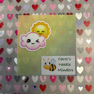 May include: A pink and white cloud with a smiling face and a winking eye, with a yellow sun with a smiling face above it. The image is on a green mesh background. A white label with a bee and the text "Carol's Needle Minders" is below the cloud.