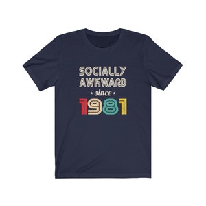 May include: Navy blue t-shirt with a vintage-style graphic that reads "Socially Awkward since 1981". The text is in white with red, yellow, green, and blue accents.