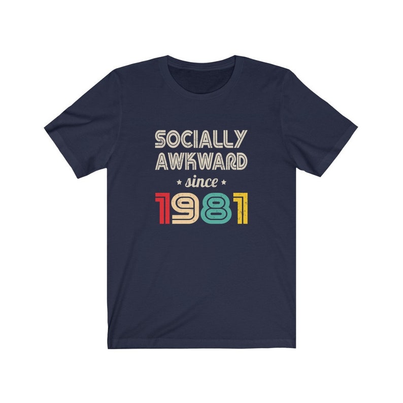 May include: Navy blue t-shirt with a vintage-style graphic that reads "Socially Awkward since 1981". The text is in white with red, yellow, green, and blue accents.