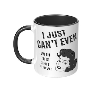 May include: White ceramic coffee mug with a black rim and handle. The mug features a black and white illustration of a woman with the text "I just can't even with this shit show!"