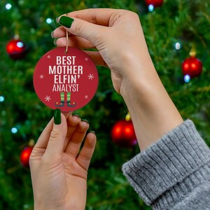 May include: Red and white Christmas ornament with the text "Best Mother Elfin' Analyst" and a cartoon elf with green shoes and pants.