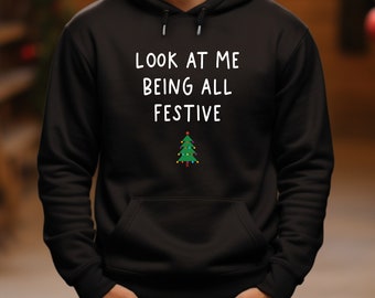 May include: Black hoodie with the white text "LOOK AT ME BEING ALL FESTIVE". A small green Christmas tree graphic with colourful lights is below the text. The person is wearing green trousers.