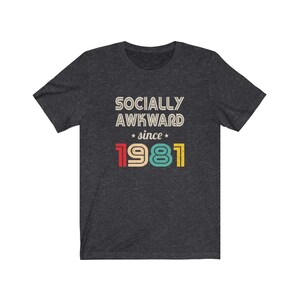 May include: Dark heather grey t-shirt with a vintage-style graphic that reads "Socially Awkward since 1981". The text is in white with red, yellow, blue, and green accents.