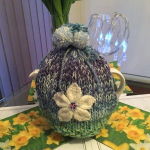 May include: A knitted teapot cosy with a white flower design and blue pom poms. The cosy is purple, blue, and green.