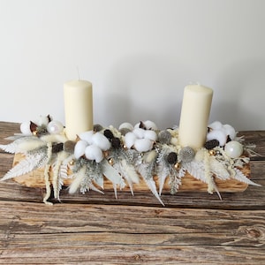 Peut inclure: Un centre de table en bois avec deux bougies blanches, des boules de coton blanches et des feuillages secs blancs et gris. Le centre de table est posé sur une surface en bois.