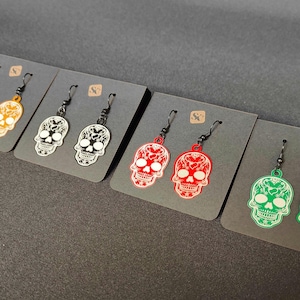 May include: A collection of skull-shaped earrings in various colors, including orange, silver, red, and green. Each earring features a detailed skull design with black hooks, displayed on gray cards.