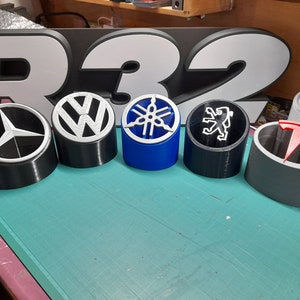 May include: Six different car logos are displayed on black, gray, and blue cylindrical bases. The logos are Mercedes-Benz, Volkswagen, Yamaha, Peugeot, and Tesla. The logos are all white except for the Tesla logo, which is red.