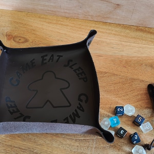May include: A brown leather dice tray with a felt lining and a black meeple design. The tray has a stitched handle and the text "Game Eat Sleep Game" around the meeple. A brown leather dice bag and a set of dice are also shown.