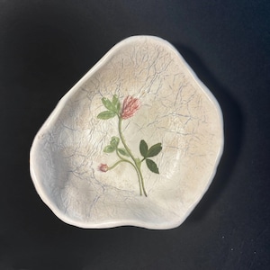 May include: An irregular, off-white ceramic bowl with a textured, crinkled surface finish. The center of the bowl features a detailed, hand-painted illustration of a pink clover flower with green leaves and stems, rendered in soft, natural tones.
