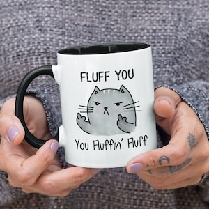 May include: White ceramic coffee mug with a black rim and handle. The mug features a cartoon cat with a grumpy expression and the text "Fluff You You Fluffin' Fluff".