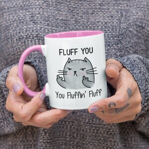 May include: White ceramic mug with a pink rim and handle. The mug features a cartoon cat with a grumpy expression giving the middle finger. The text on the mug reads "Fluff You You Fluffin' Fluff".