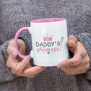 May include: White ceramic mug with a pink rim and handle. The mug has a pink crown and the text "Daddy's princess" in black and green.