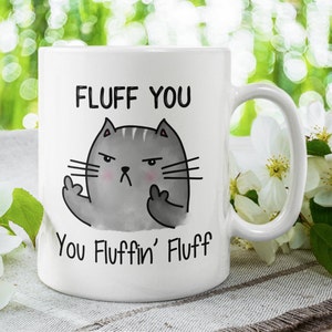 May include: White ceramic mug with a gray cat illustration giving the middle finger. The text on the mug reads "Fluff You You Fluffin' Fluff".