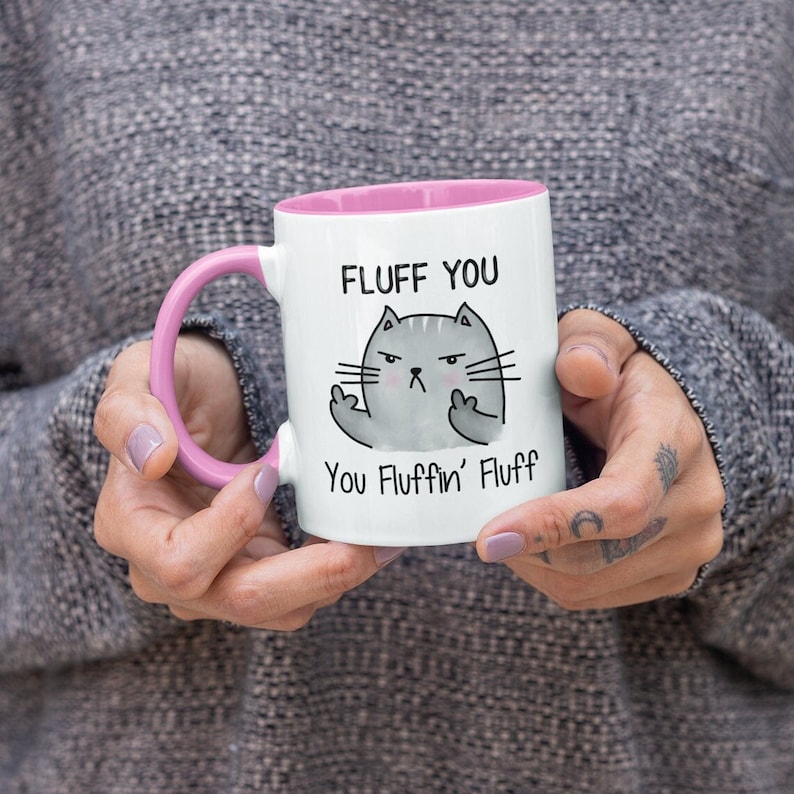 May include: White ceramic mug with a pink rim and handle. The mug features a cartoon cat with a grumpy expression giving the middle finger. The text on the mug reads "Fluff You You Fluffin' Fluff".