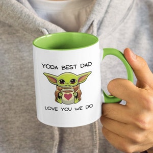 May include: White ceramic mug with a green rim and handle. The mug features a cartoon image of a green alien character holding a cup of coffee with a red heart on it. The text "YODA BEST DAD LOVE YOU WE DO" is printed on the mug.