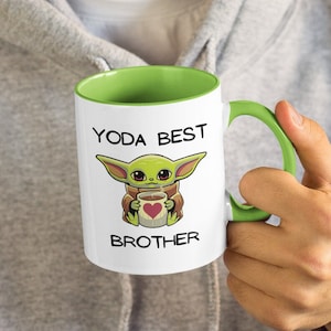 May include: White ceramic mug with a green rim and handle. The mug features a cartoon image of a green alien character holding a cup with a red heart. The text "YODA BEST BROTHER" is printed on the mug.