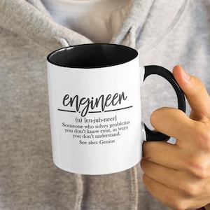 Op de afbeelding: Een witte keramische mok met een zwarte rand en handvat. De mok heeft een zwart-wit ontwerp met het woord "engineer" en een definitie van het woord. De definitie luidt: "(n) [en-juh-neer] Iemand die problemen oplost waarvan je niet weet dat ze bestaan, op manieren die je niet begrijpt. Zie ook: Genius."
