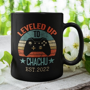 以下が含まれることがあります： 「LEVELED UP TO CHACHU EST. 2022」とレトロなビデオゲームコントローラーのグラフィックが描かれた白いデザインの黒いコーヒーカップです。