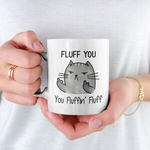 May include: White ceramic coffee mug with a black and gray cartoon cat giving the middle finger. The mug reads "Fluff You You Fluffin' Fluff".
