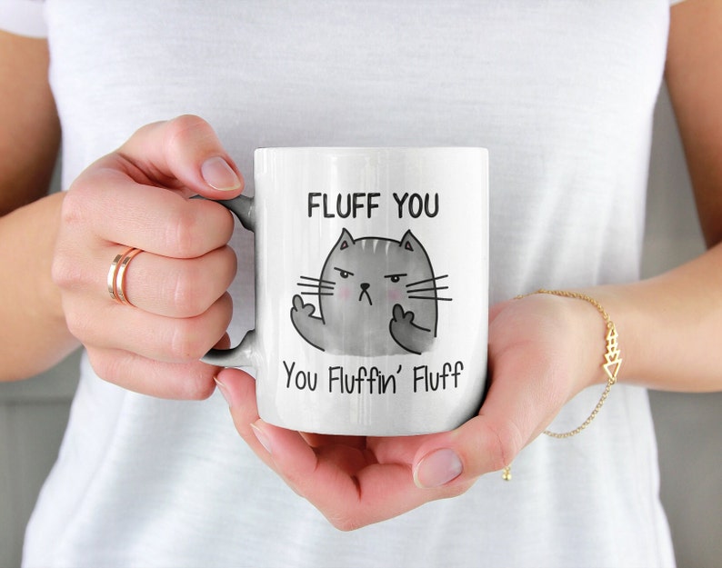 May include: White ceramic coffee mug with a black and gray cartoon cat giving the middle finger. The mug reads "Fluff You You Fluffin' Fluff".