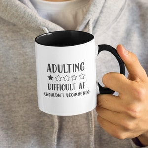 May include: White coffee mug with a black interior and handle. The mug has black text that reads "Adulting  ★★★★★ Difficult AF (Wouldn't Recommend)"